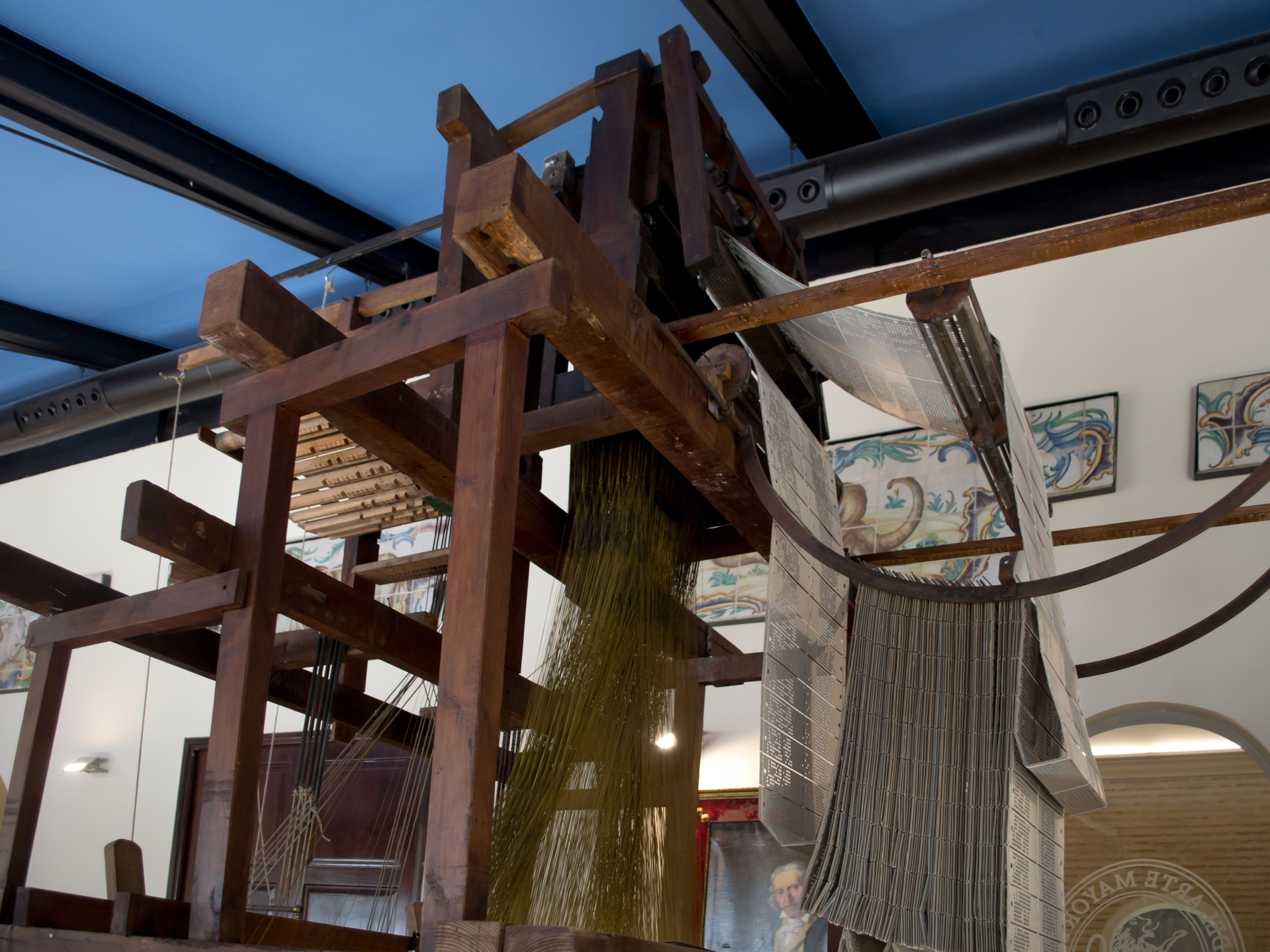 A wooden Jacquard attachment stands over a handloom, with laced cards passing around its upper mechanism and harness cords hanging below.