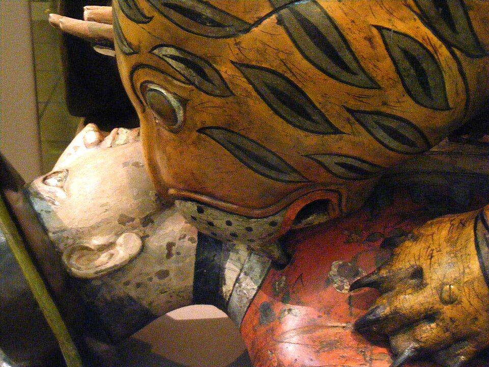 Close view of the wooden tiger’s head above the European figure’s face and red coat.