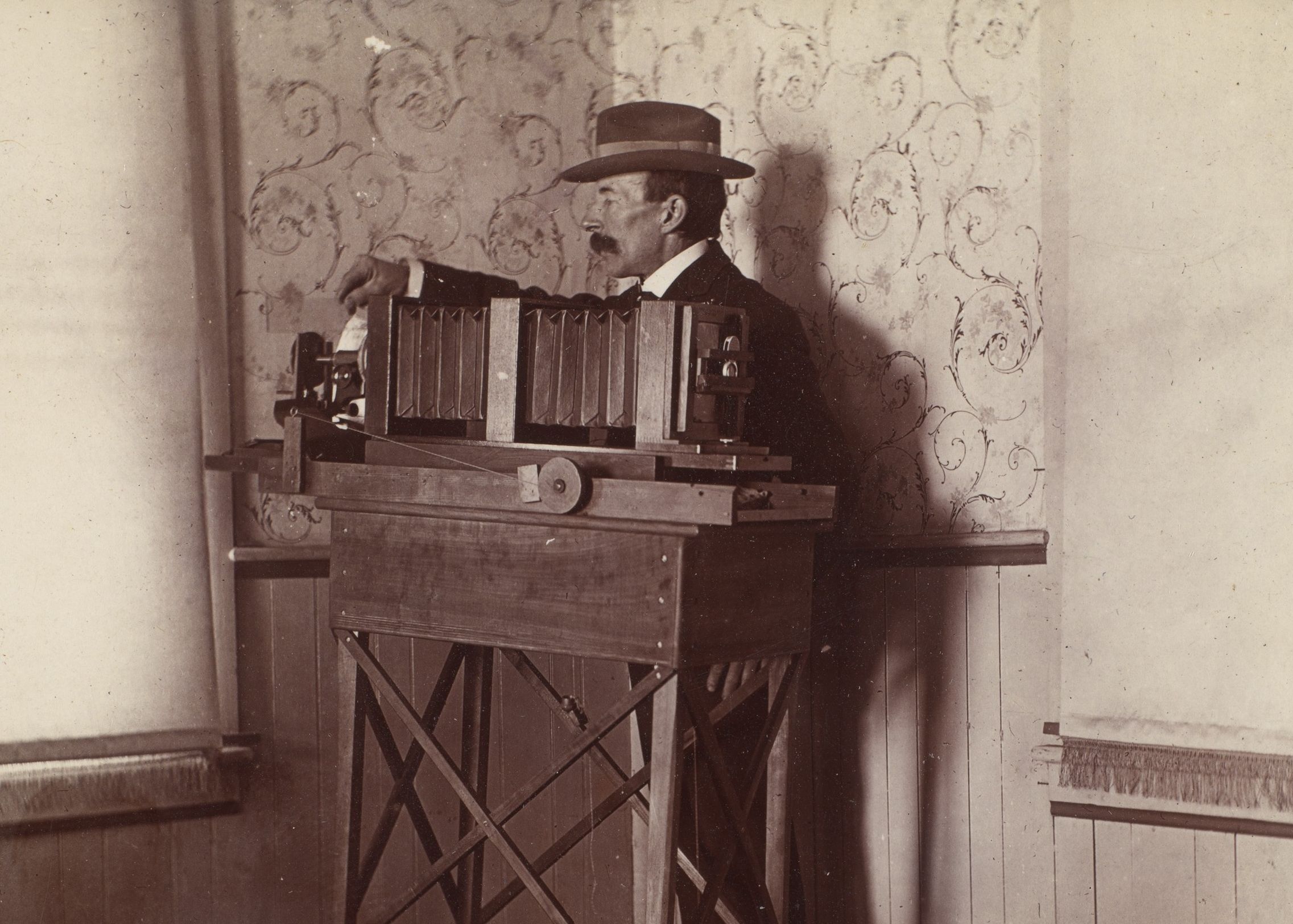 Wilson Bentley standing behind a long bellows camera mounted on a wooden table beside a window.
