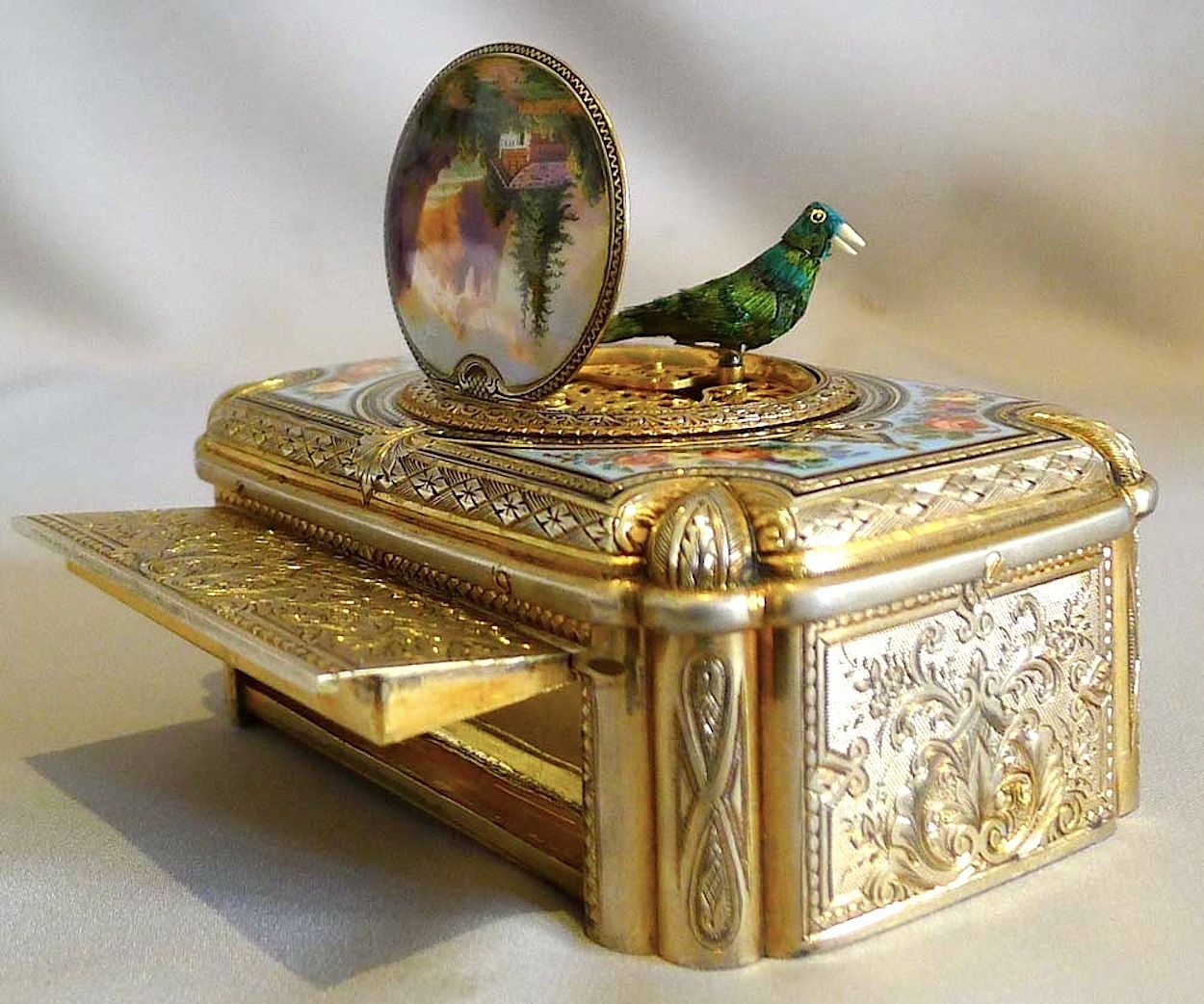 A green feathered mechanical bird stands above an open silver-gilt box, beside its raised enamel lid and open tobacco compartment