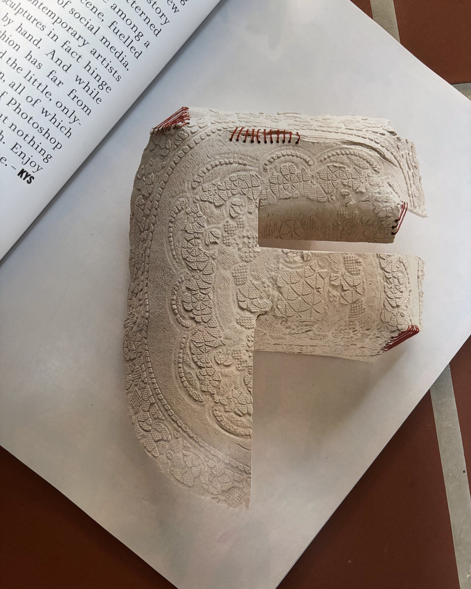 Porcelain letter F, its surface imprinted with lace and edged with hand-stitched red thread, resting on an open magazine over terracotta tiles.