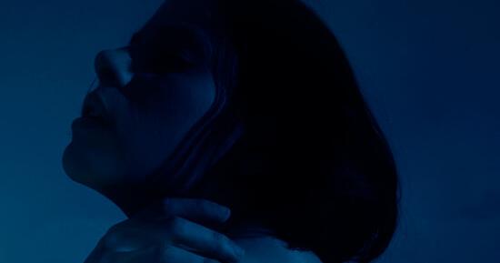 Woman with short hair, head tilted back, and hand on her neck, illuminated by moody blue and teal light.