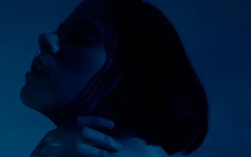 Woman with short hair, head tilted back, and hand on her neck, illuminated by moody blue and teal light.