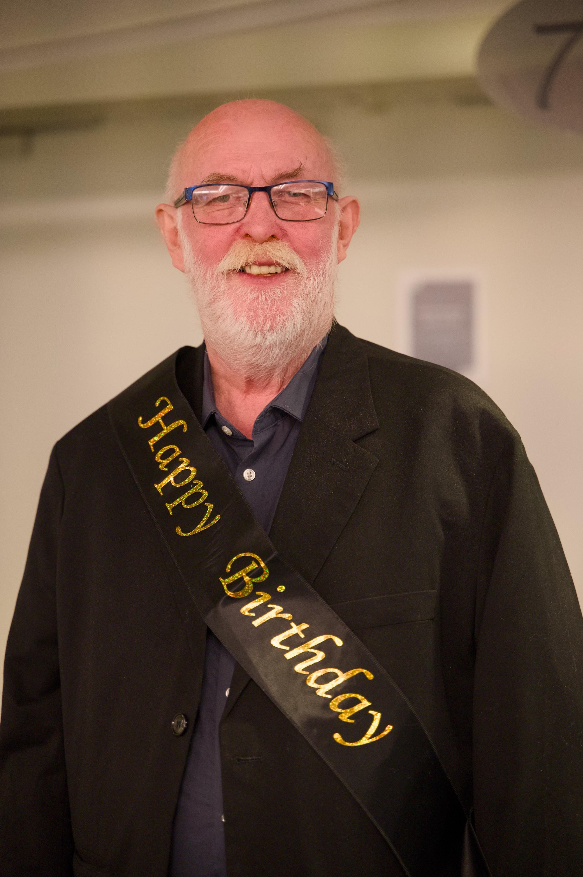 Alf Steinar smiler med eit svart band med gullskrift der det står "gratulerer med dagen" på engelsk.