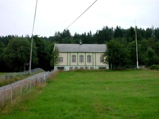 Bilete av lysegult ungdomshus meg grøn skog i bakgrunnen. Foran er det grønt gras og heilt til venstre i bilete er det ein grusveg som går mot huset.