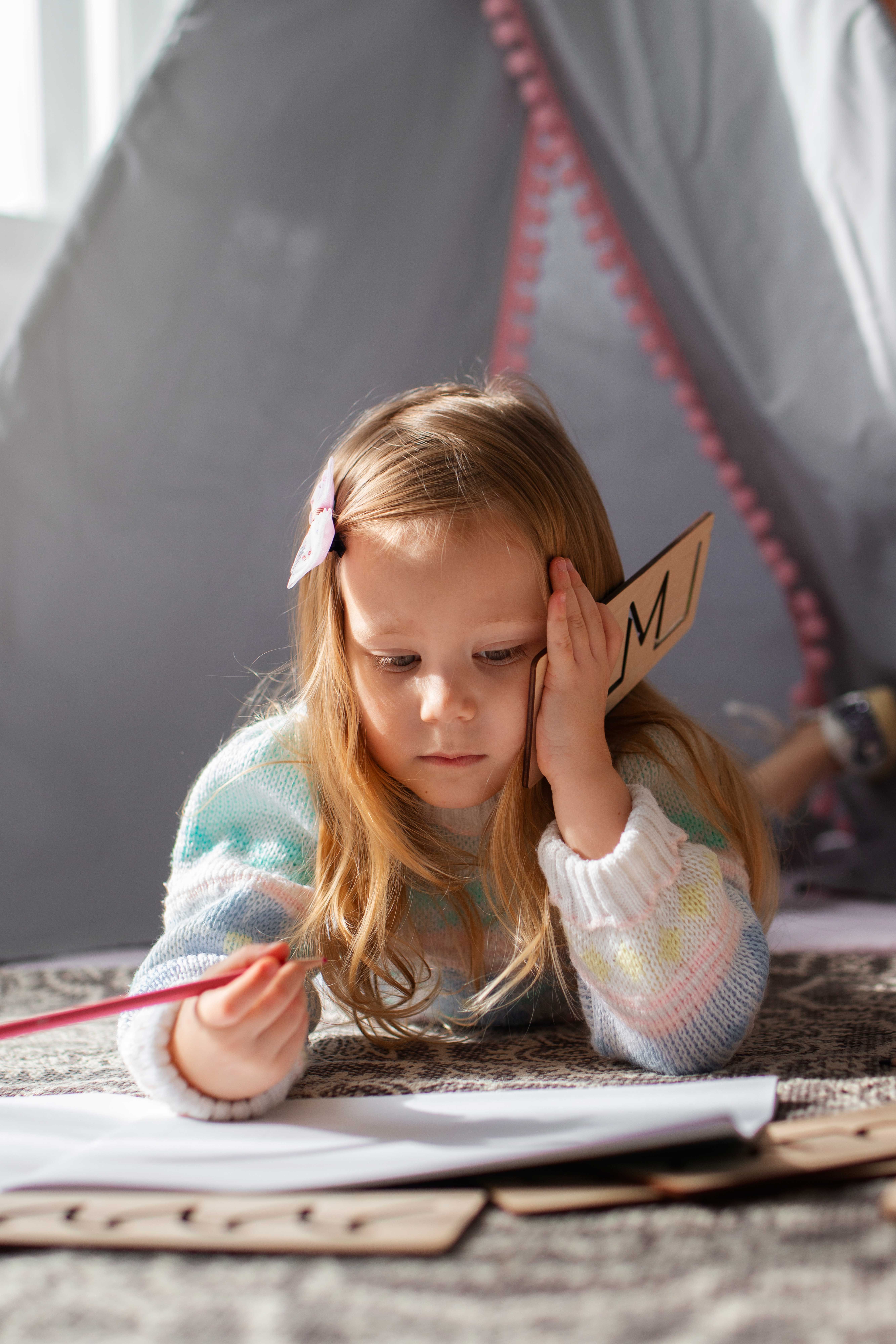 Fillette allongée, concentrée sur son cahier, tenant une lettre en bois, illustrant les difficultés d’apprentissage liées à la dyslexie.