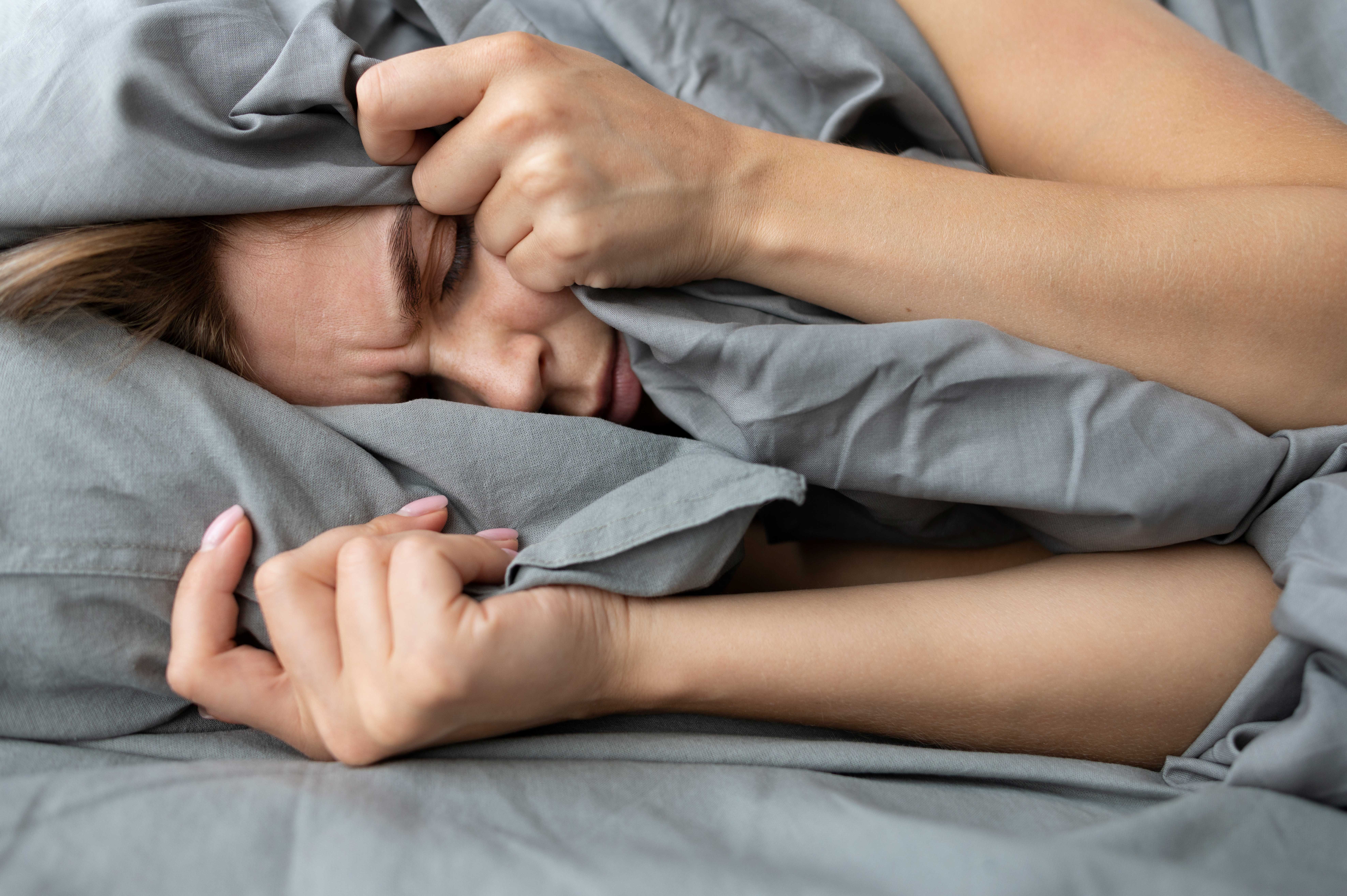 Femme allongée dans un lit, visiblement agitée et en proie à l'insomnie, serrant sa couverture avec une expression de frustration, symbolisant les difficultés liées aux troubles du sommeil