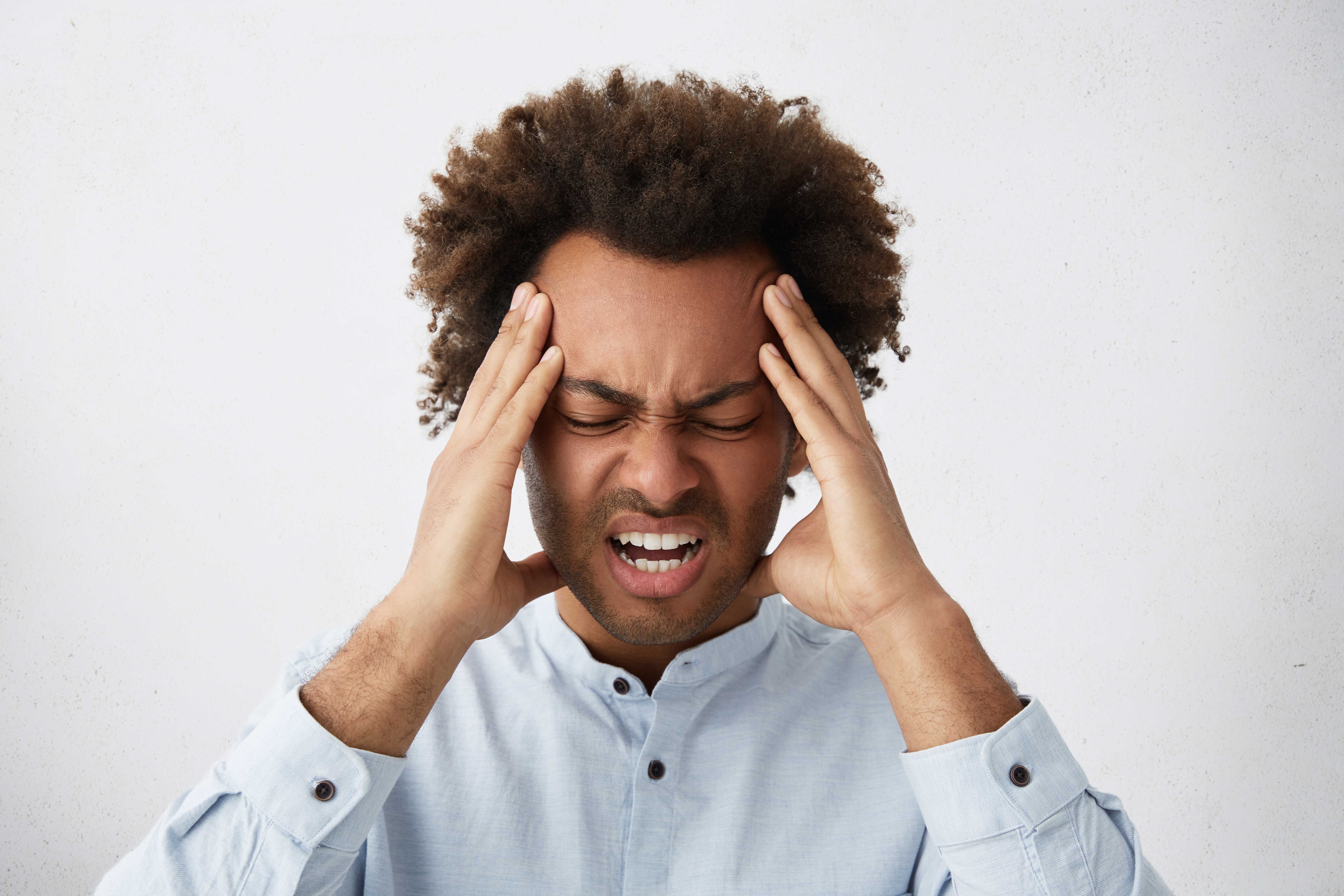 Homme, les yeux fermés, tenant sa tête des deux mains, exprimant une douleur ou une tension liée au stress. L'arrière-plan est clair, soulignant le focus sur son expression tendue et accablée.