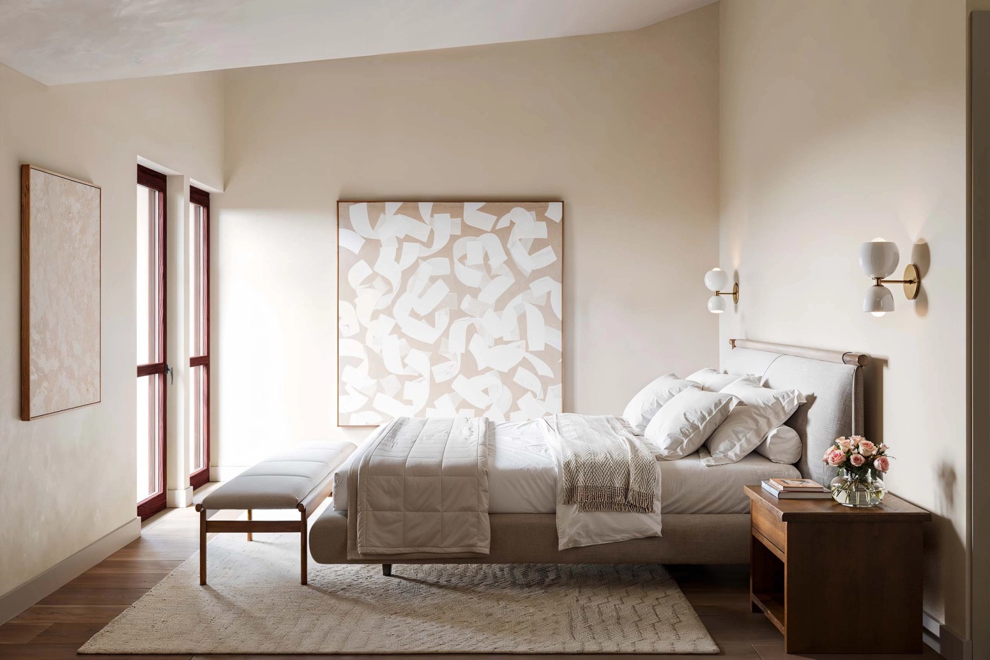 Light bedroom with an abstract artwork, wall lamps and a bench at the foot of the bed.