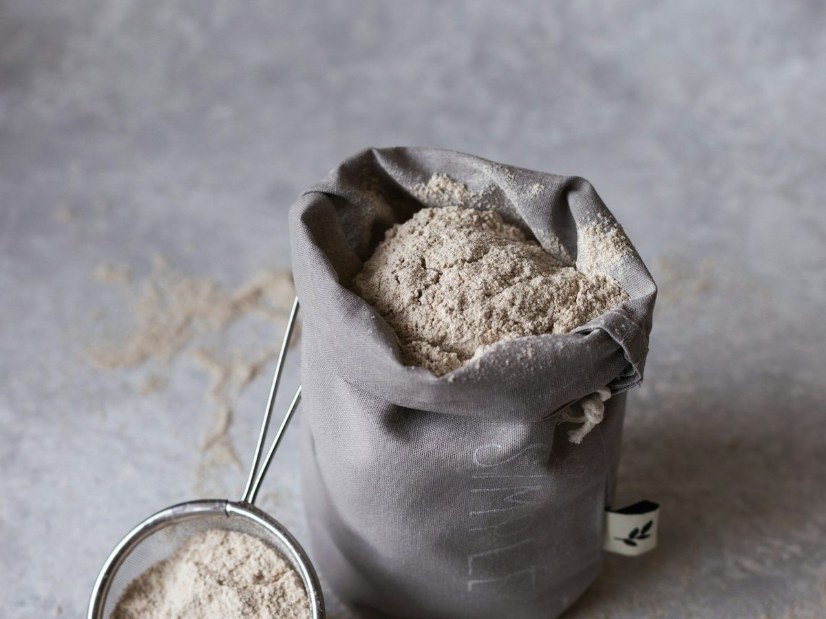 A cloth bag of flour next to a small sieve on a grey surface.