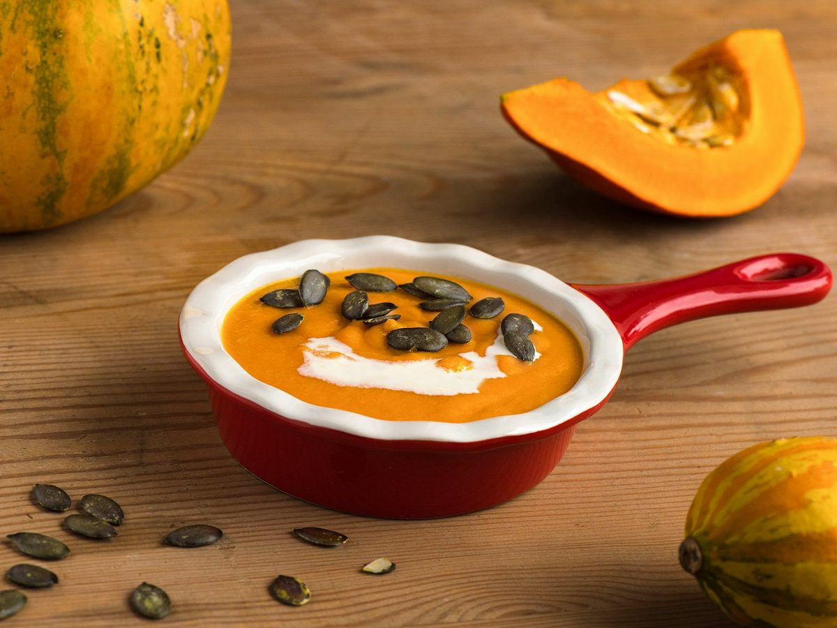 A small red dish of smooth orange pumpkin soup with a swirl of cream and pumpkin seeds, beside squash on a wooden table.