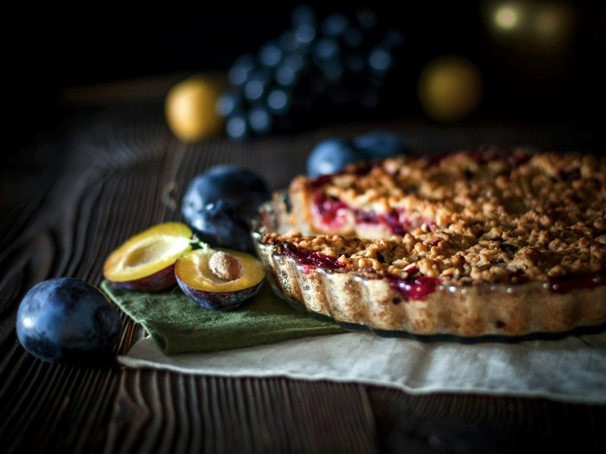 A plum tart with a crumbly streusel topping on a wooden table, with halved purple plums beside it.