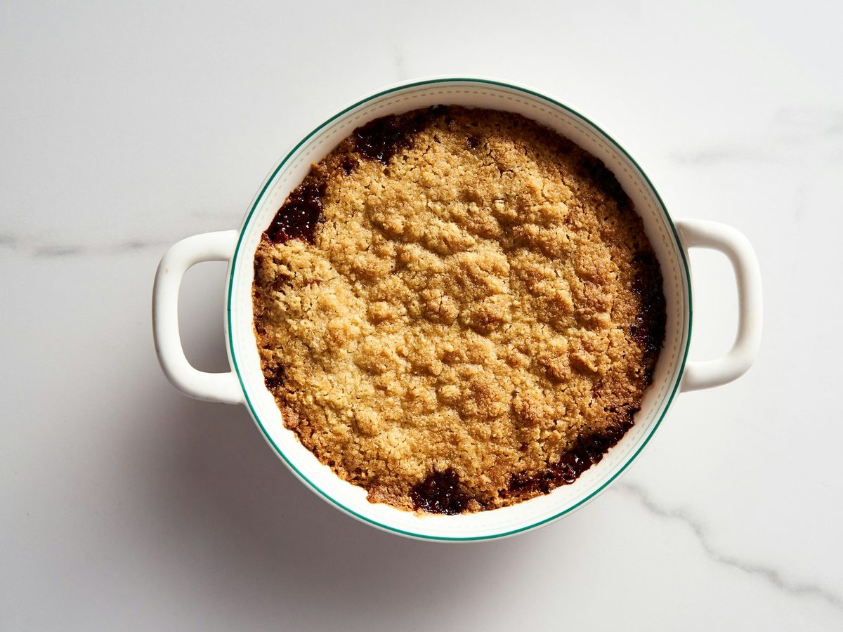 A golden crumble with a crisp, sandy topping in a round white baking dish with handles, on a white marble counter.