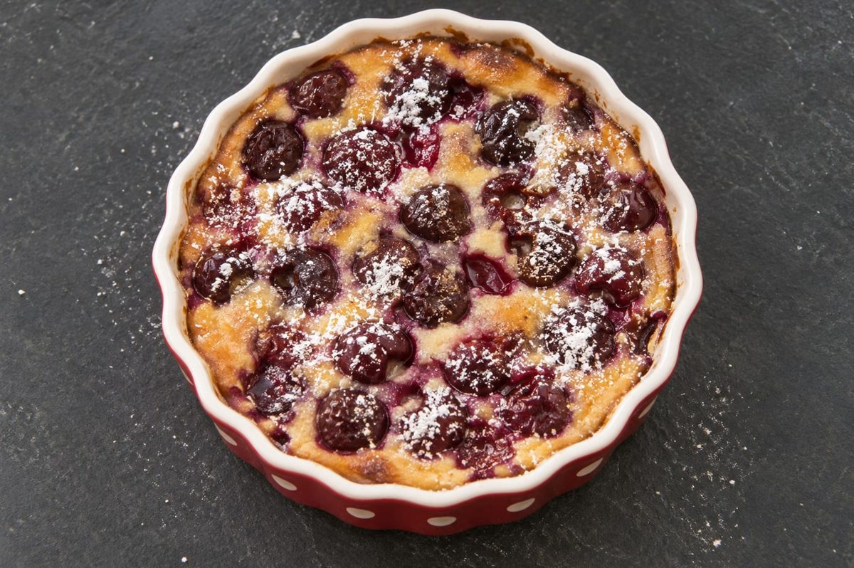 A cherry clafoutis dusted with icing sugar in a red fluted baking dish on dark slate.