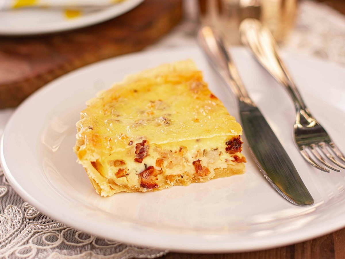 A slice of quiche lorraine with pieces of bacon in a golden custard, on a white plate with a knife and fork.