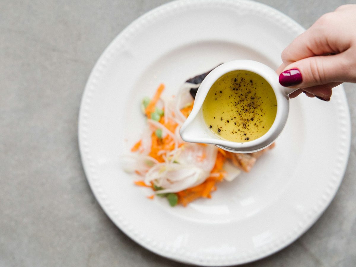 A hand pouring mustard vinaigrette with black pepper from a small jug onto a salad.