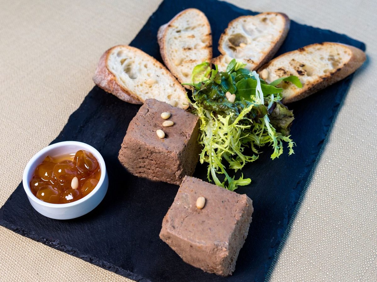Two thick slices of country terrine on a slate with toasted bread, salad and a small dish of pickles.