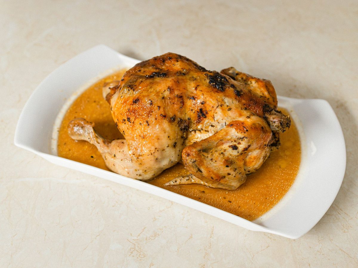 A whole golden roast chicken sitting in its pan juices on a white serving dish.