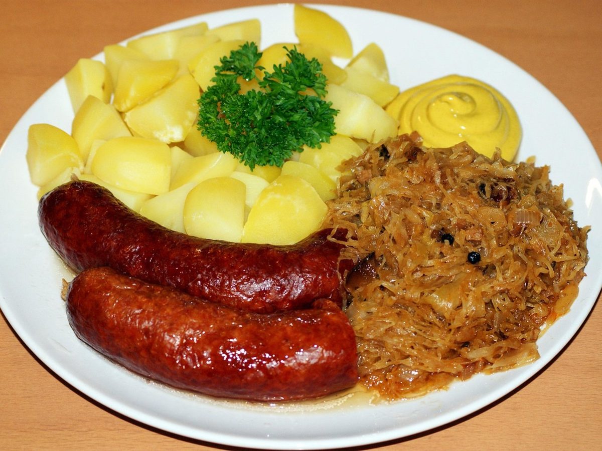 A plate of sauerkraut with two smoked sausages, boiled potatoes and a spoon of mustard.