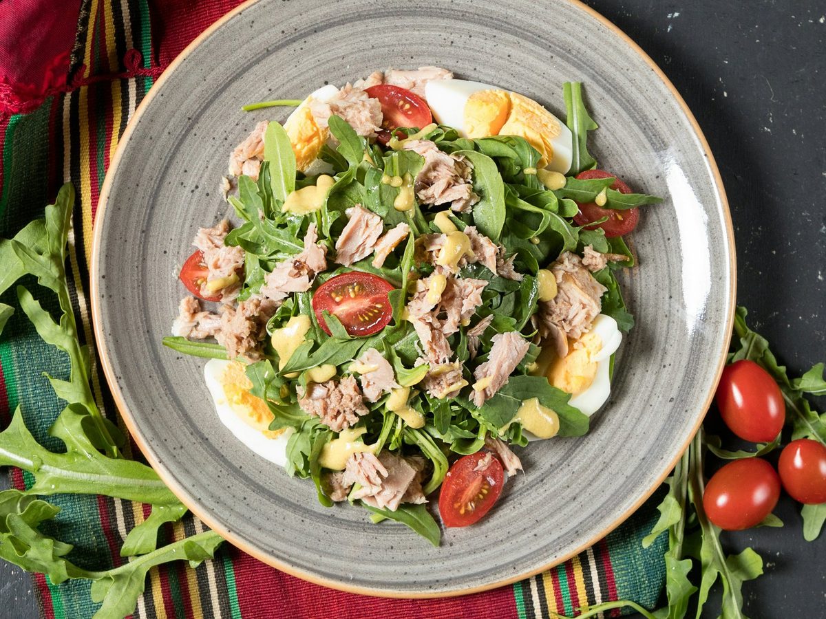 Flaked tuna, hard-boiled egg and halved cherry tomatoes on arugula with a mustard dressing, on a grey plate.