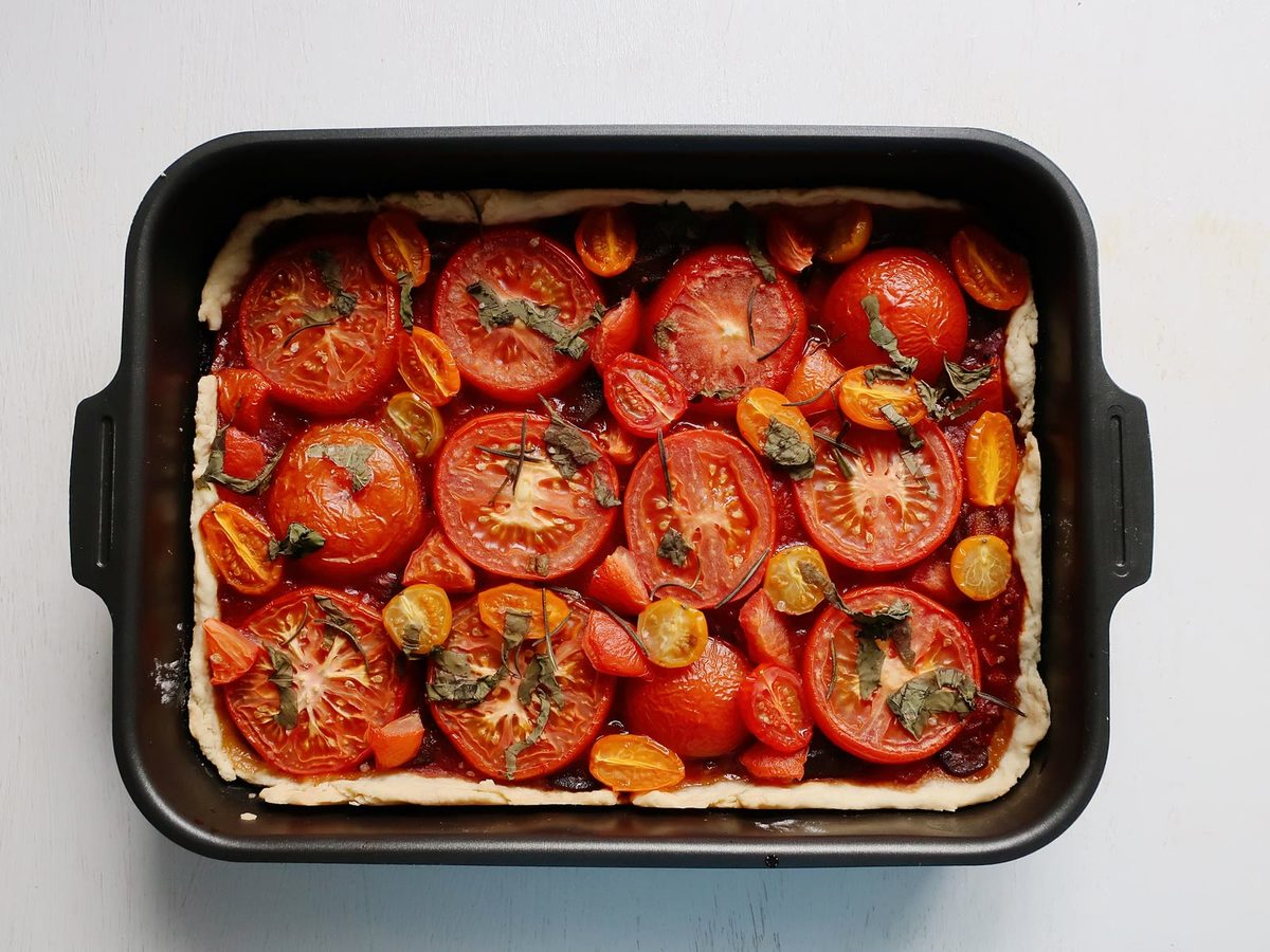 A rectangular tomato tart with sliced red and yellow tomatoes and herbs on a pale crust, in a black baking dish.