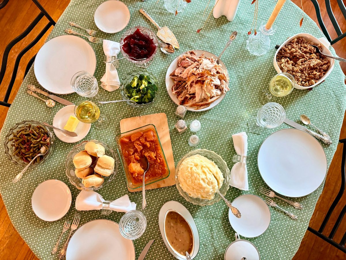 A home table set for an autumn holiday dinner, seen from above, with sliced roast turkey, mashed potatoes, green beans, cranberry sauce and bread rolls.