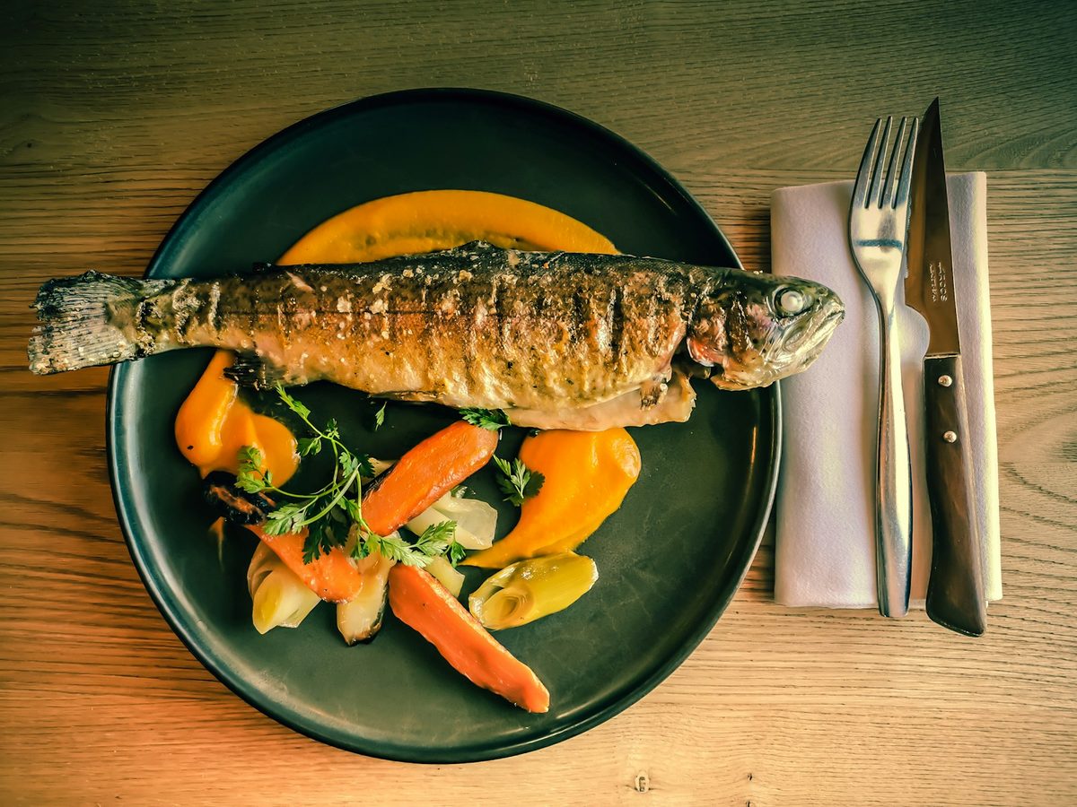 A whole pan-fried trout on a dark green plate with glazed carrots and parsley.