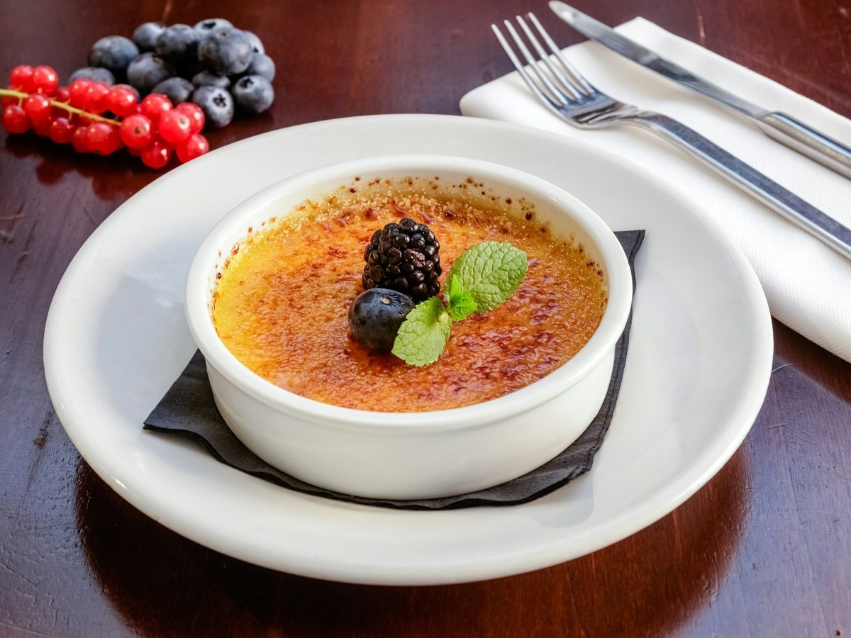 A crème brûlée in a white ramekin with berries and mint, on a plate.
