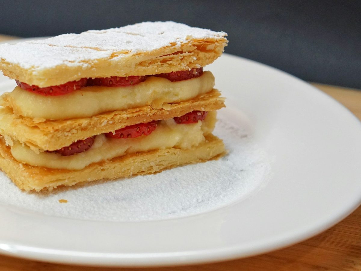 A slice of mille-feuille with layers of puff pastry, cream and strawberries on a white plate.