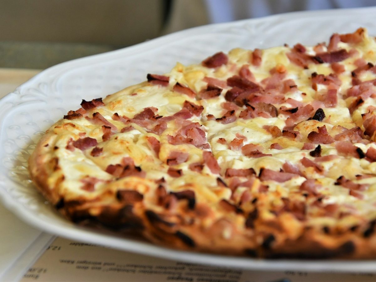 A tarte flambée with a blistered crust, covered in cream and pieces of smoked bacon, on a white plate.