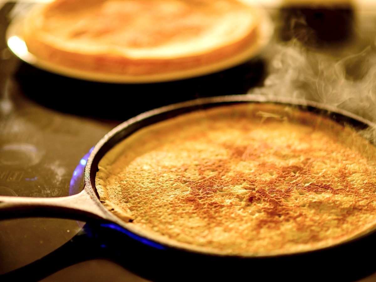 A crêpe cooking in a black steel pan, with a stack of crêpes behind it.