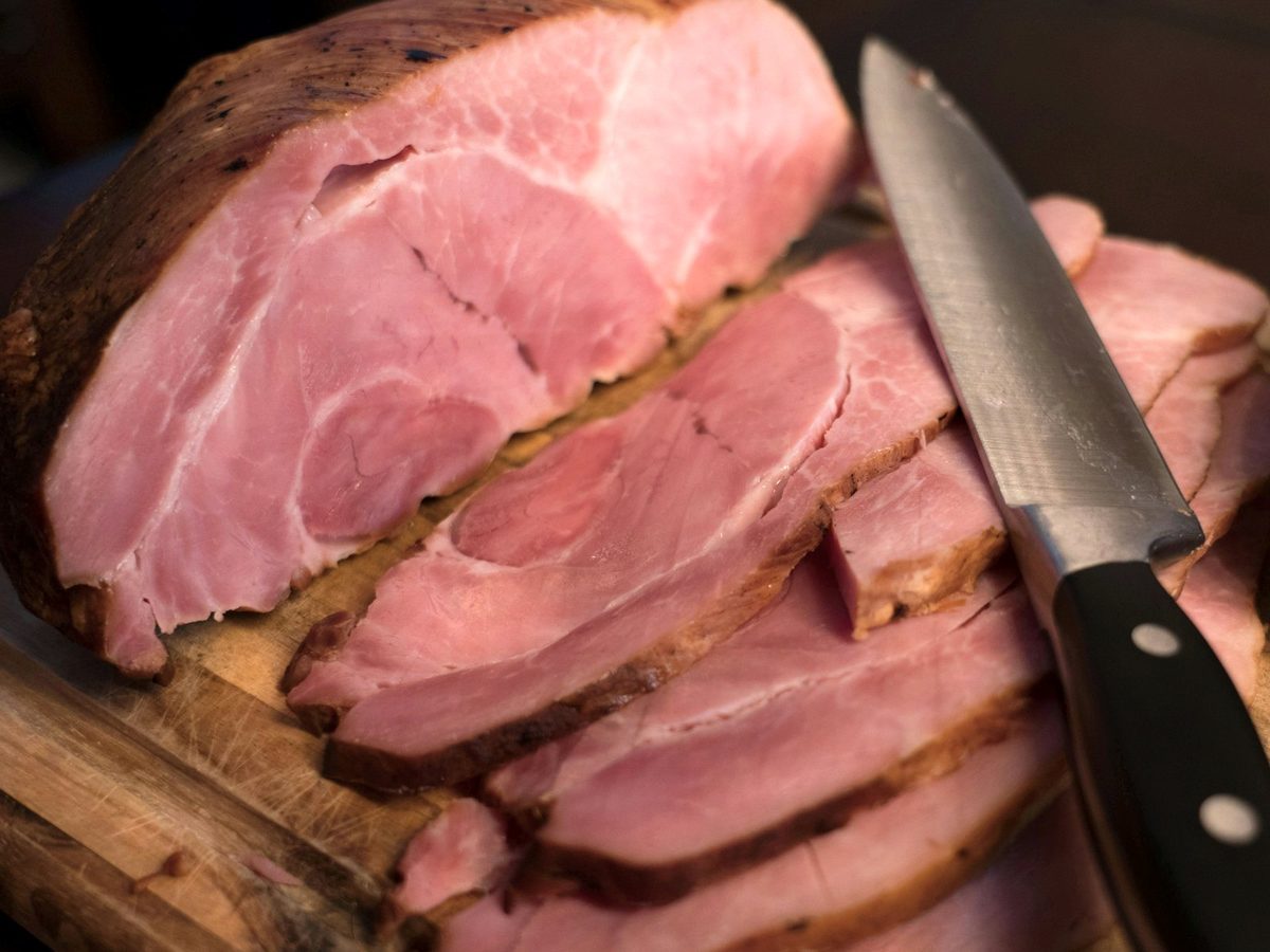 A cooked ham being sliced on a wooden board with a long knife.