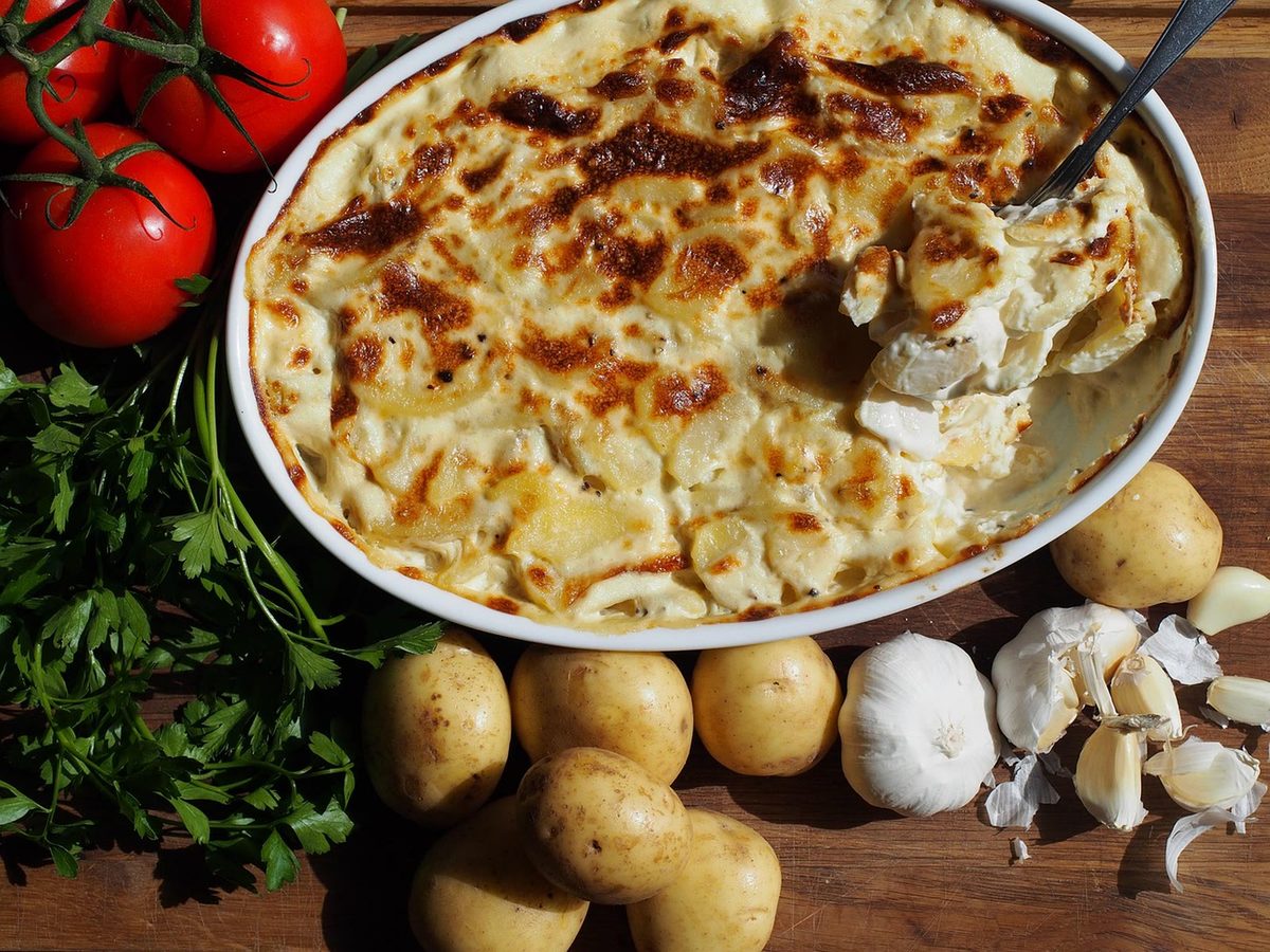 A creamy potato gratin with a browned top in an oval white dish, surrounded by potatoes, garlic, parsley and tomatoes on a wooden board.