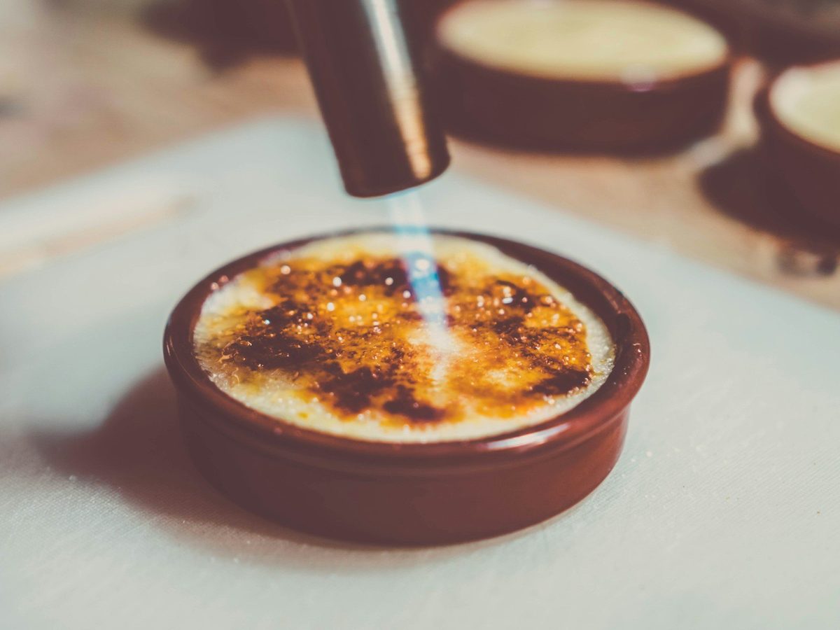 A kitchen torch caramelizing the sugar on a crème brûlée in a brown ramekin.