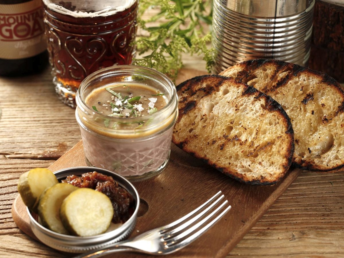 A small jar of pork rillettes on a wooden board with grilled bread slices and pickles.