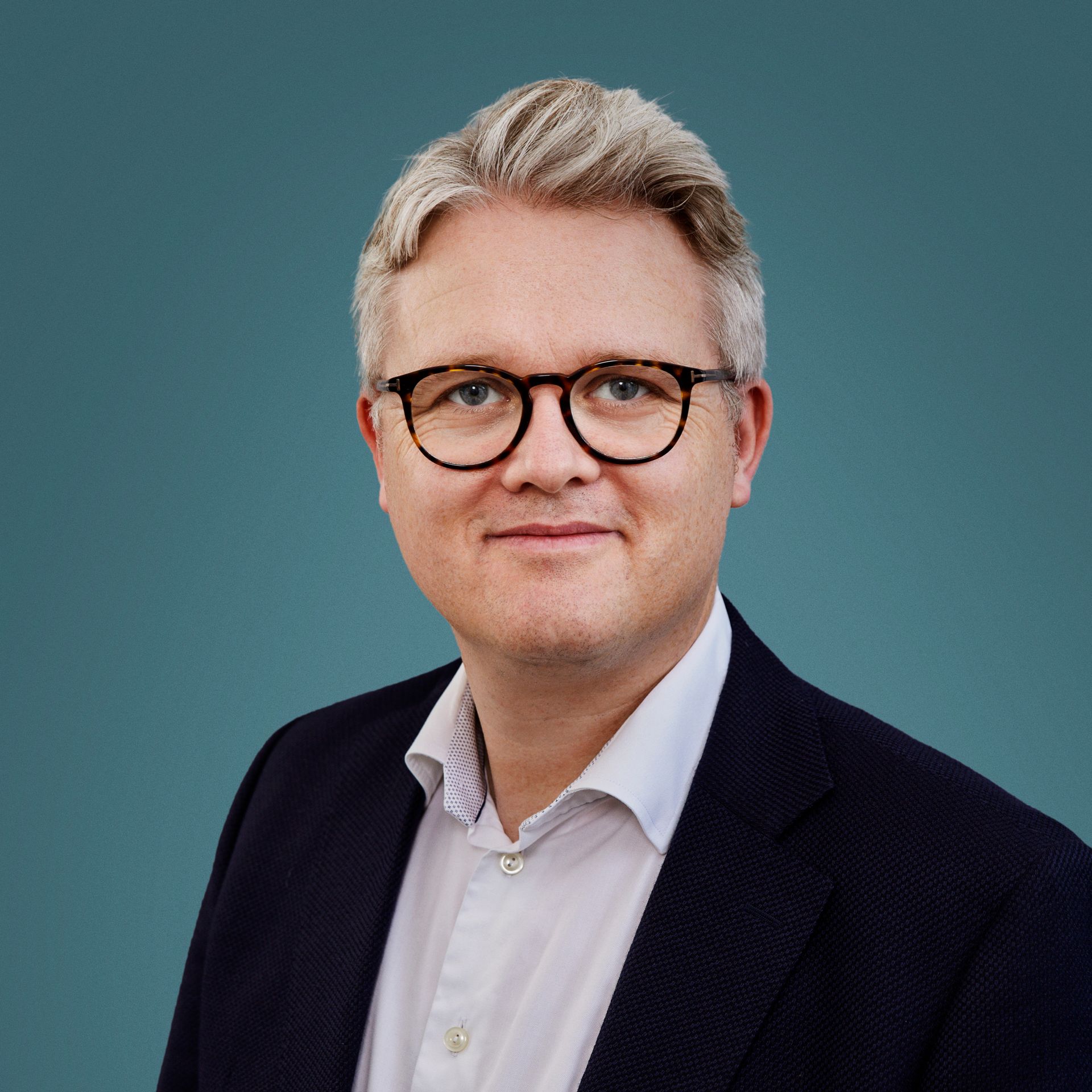 Preview of: Portræt af en mand med gråt hår og briller, iført mørk blazer og hvid skjorte mod en blågrøn baggrund.