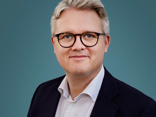 Portræt af en mand med gråt hår og briller, iført mørk blazer og hvid skjorte mod en blågrøn baggrund.