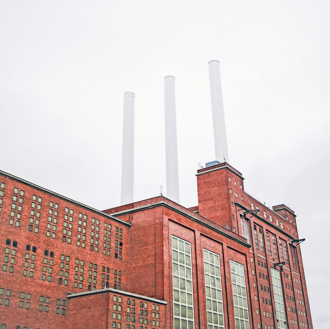 Rød industribygning i mursten med tre høje, hvide skorstene under en overskyet himmel