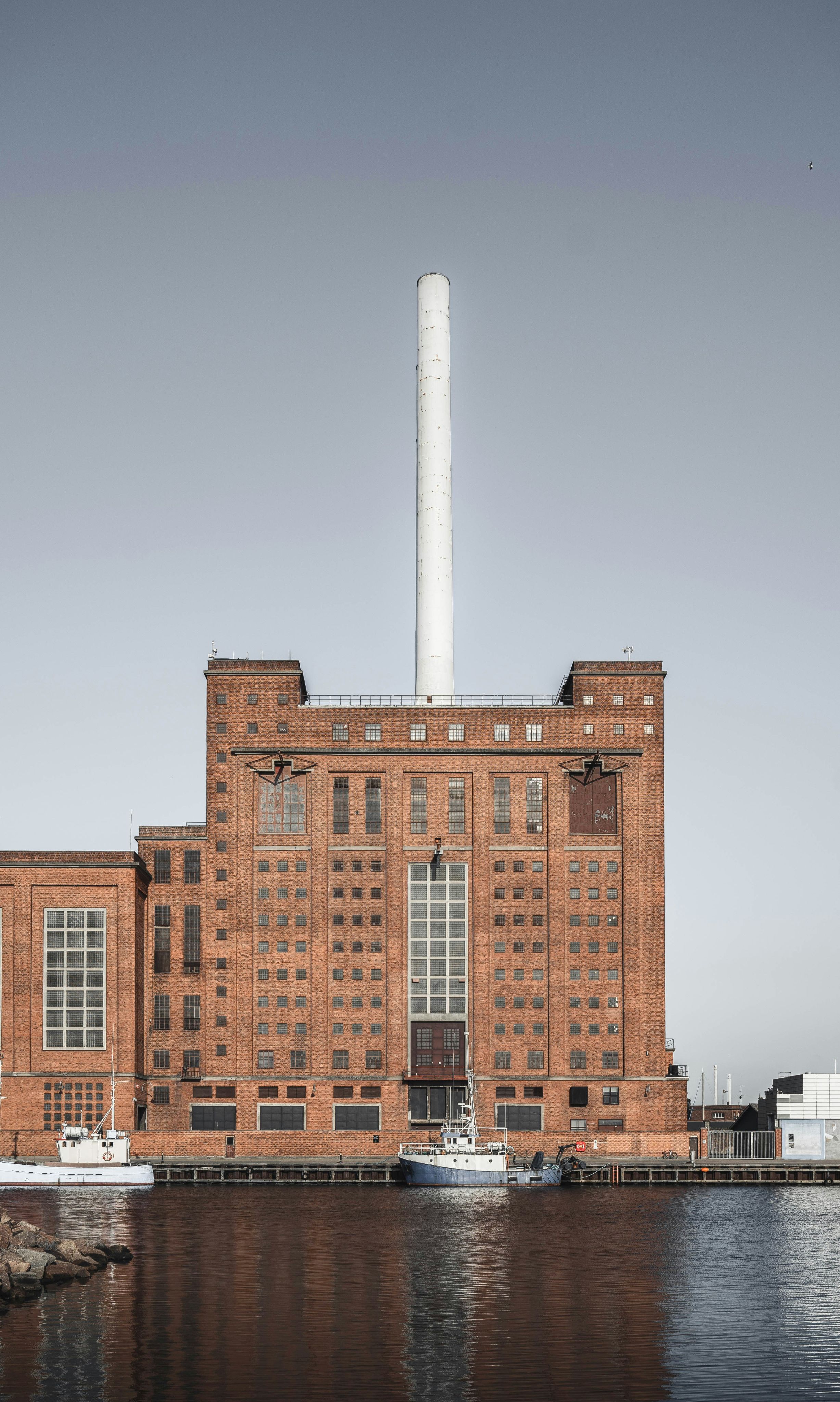 Preview of: Stor industribygning i røde mursten med hvid skorsten og både ved kajen