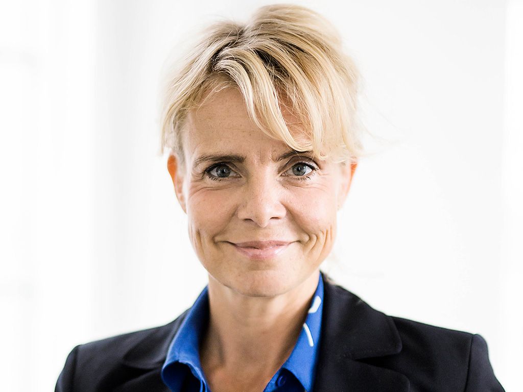 Preview of: Smilende kvinde med kort blondt hår, blå skjorte og sort blazer.