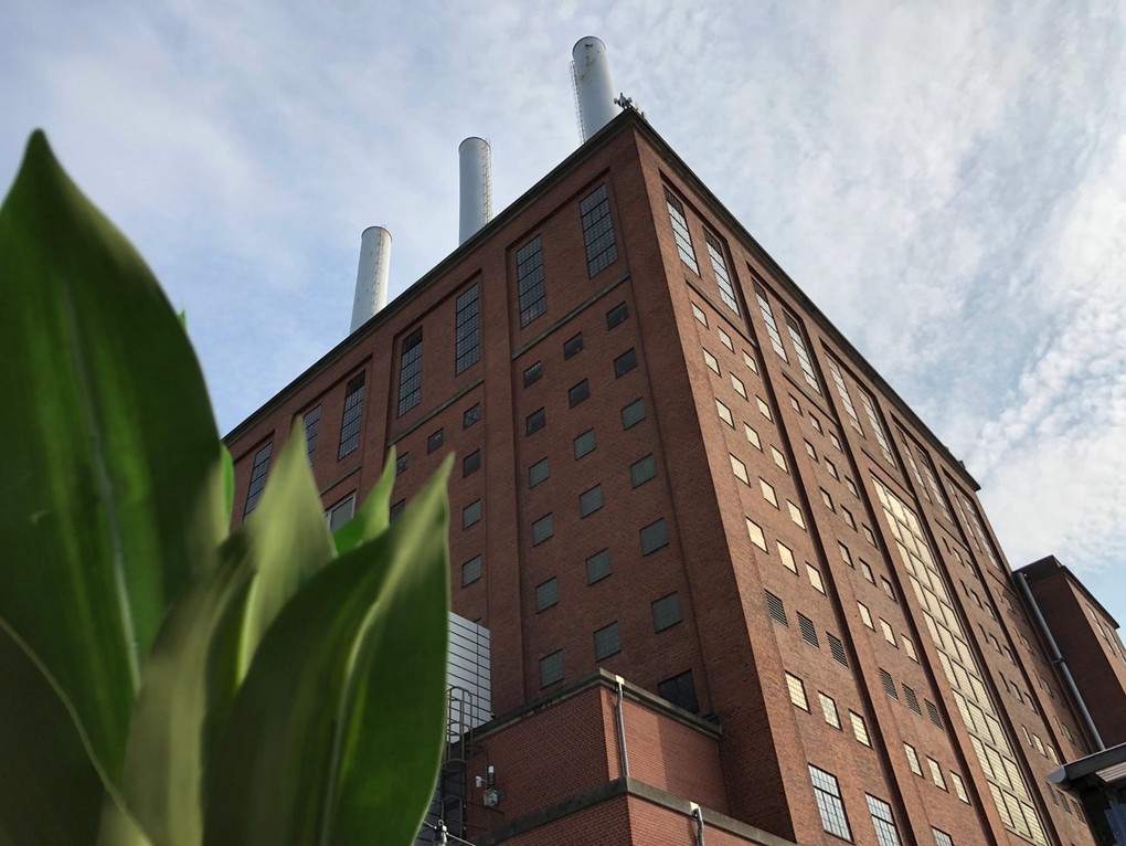 Høj industribygning i røde mursten med skorstene og grønne blade i forgrunden