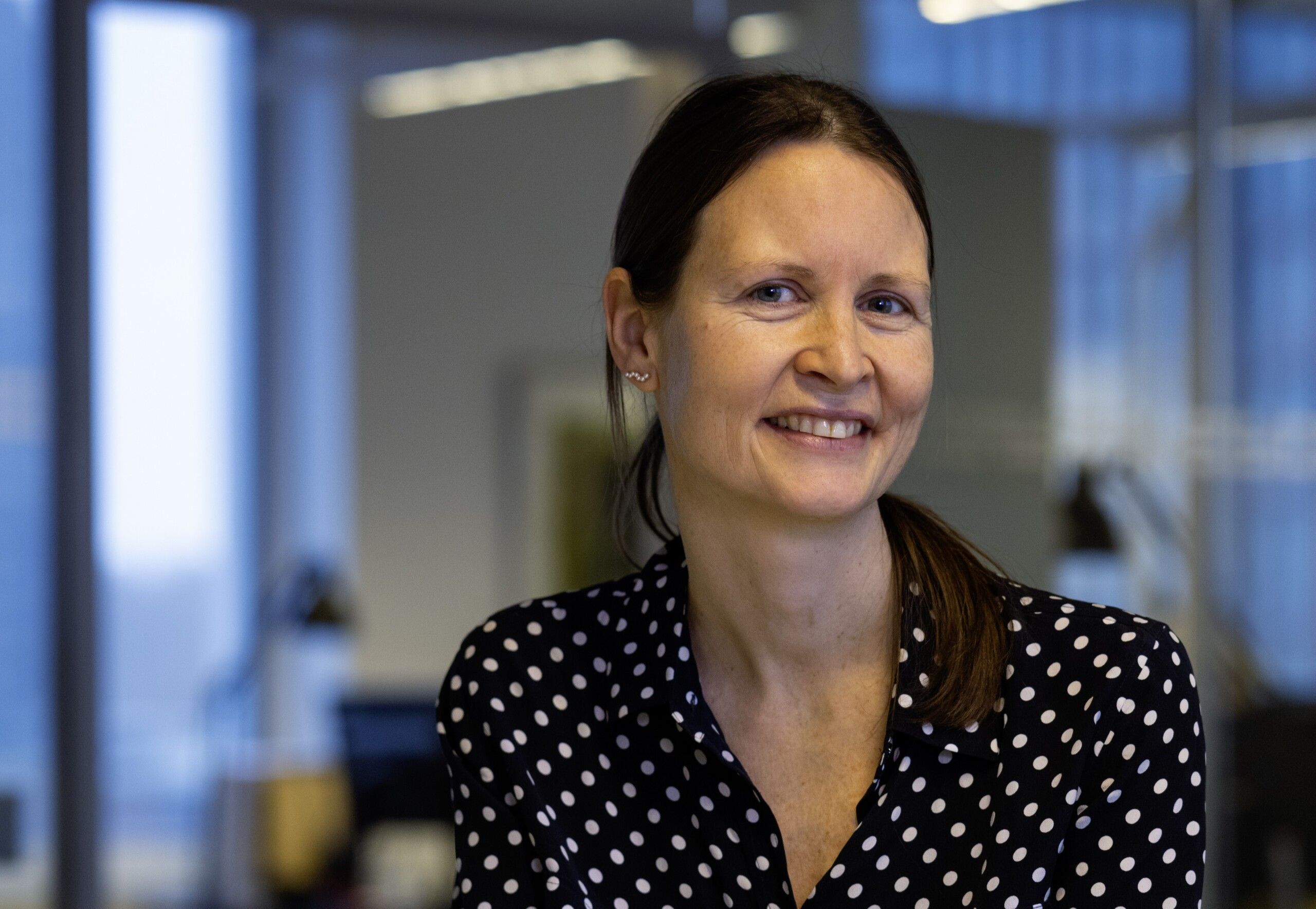 Preview of: Smilende kvinde med mørkt hår og sort, hvidprikket bluse på et kontor