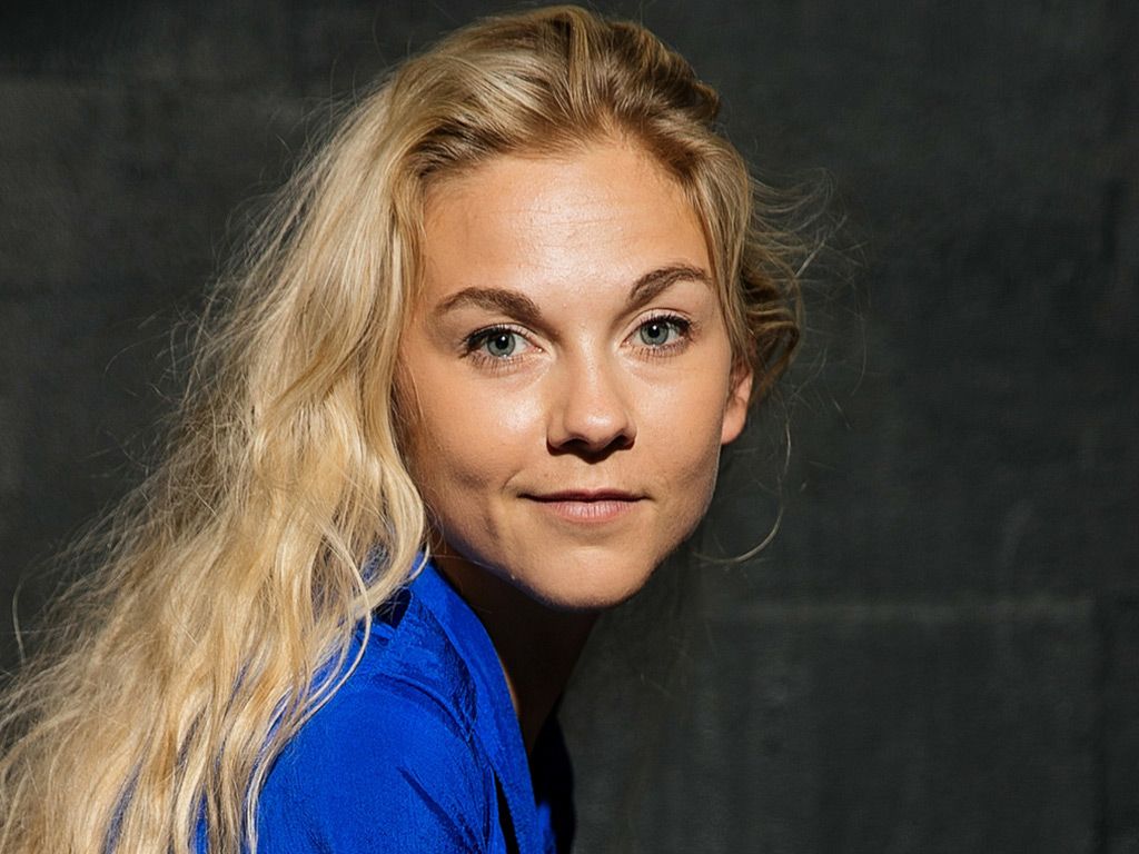 Preview of: Portræt af en blond kvinde i blå skjorte foran en mørk baggrund