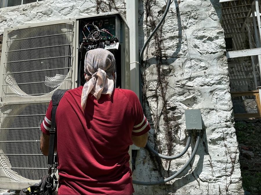 Technician working on a multi-zone heat pump outdoor unit with the service panel removed, beside a stone wall in New York