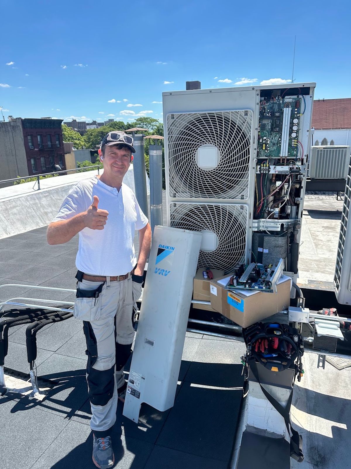 Art HVAC technician giving a thumbs-up beside a Daikin outdoor unit after an installation on a Brooklyn roof