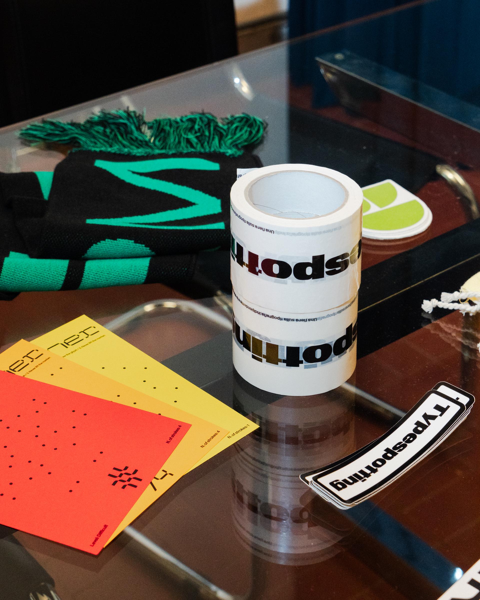 Folded green-and-black scarf, printed tape and a Typespotting sticker on a glass table.