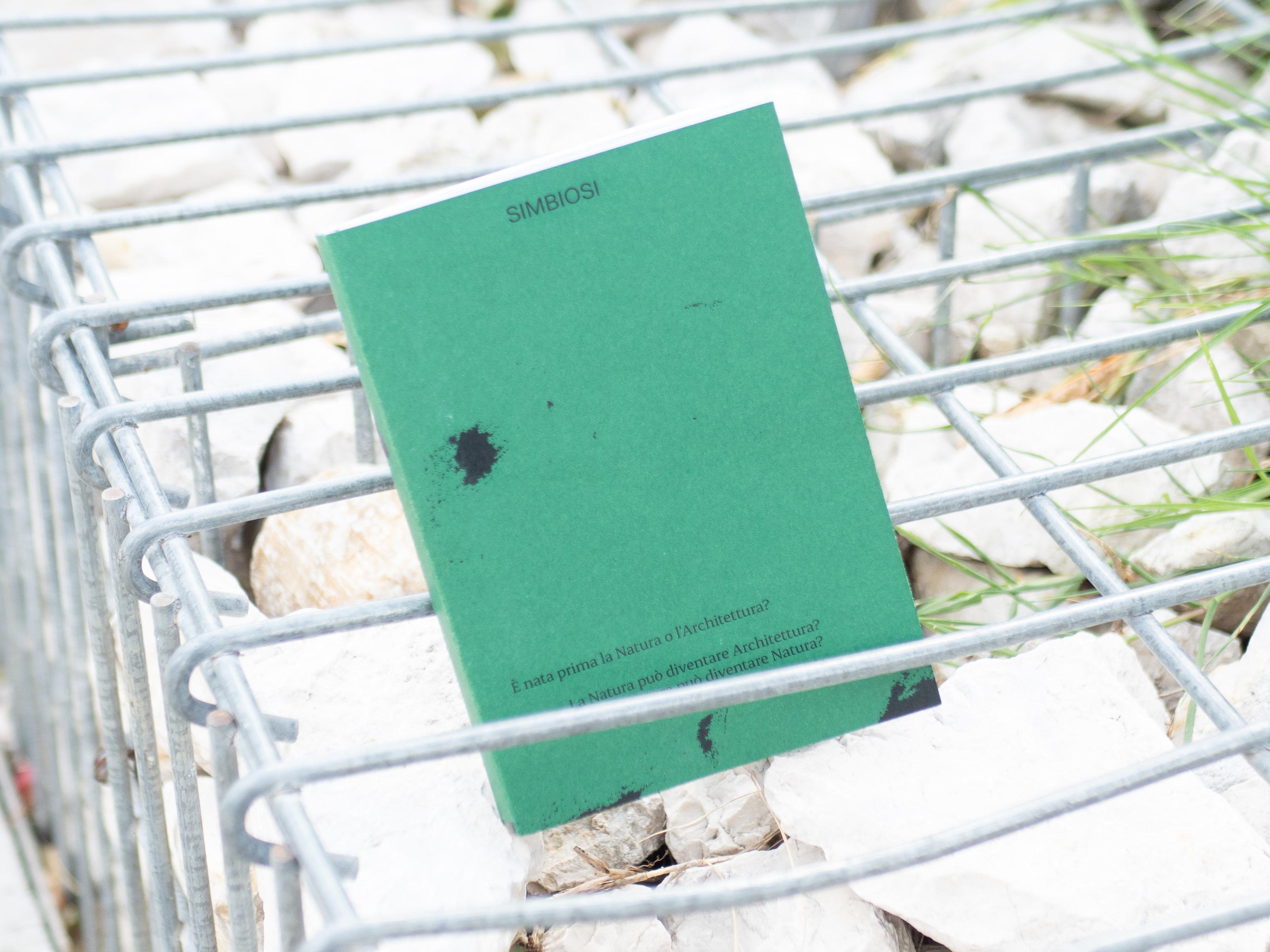 Green Simbiosi book resting on a metal grid and stones.