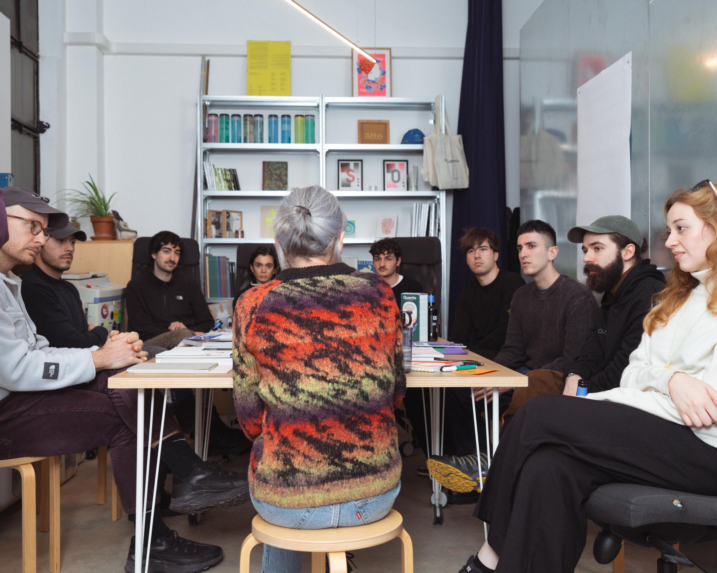 Group seated around a table in discussion, viewed behind a person in a patterned knit top.