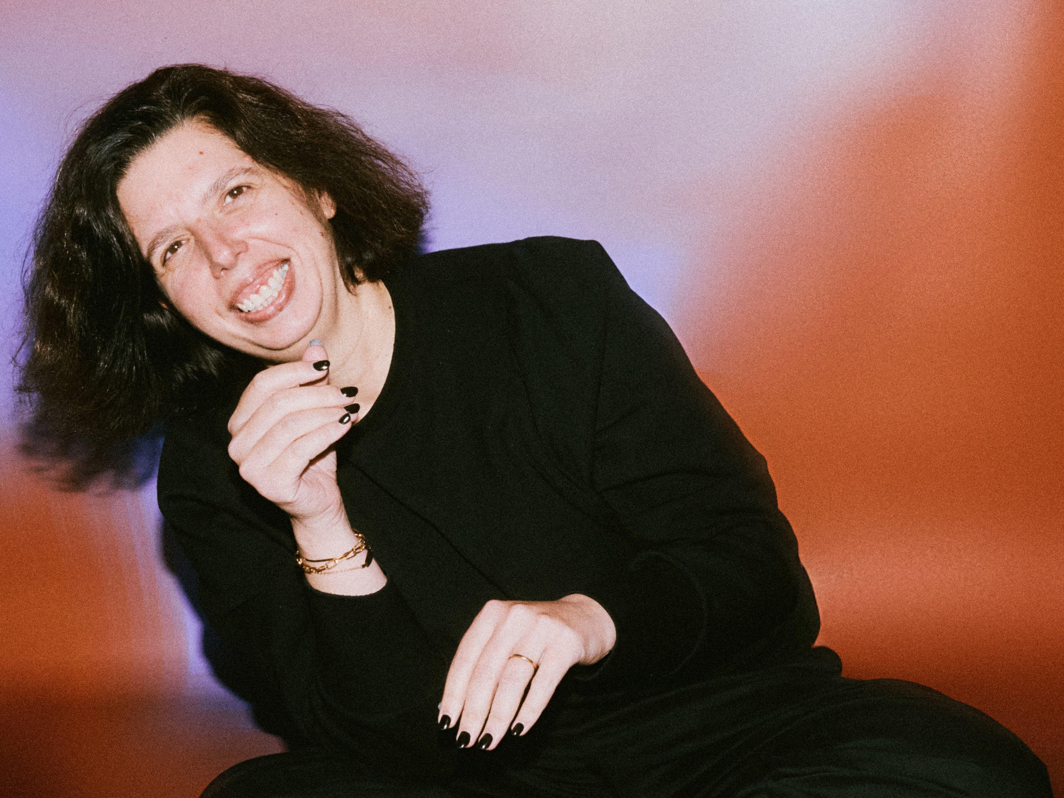 Smiling person in black leaning forward against a pink and blue background.