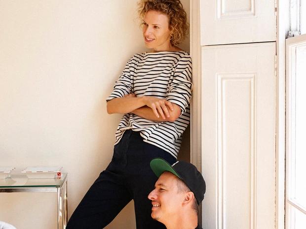 Person in a striped shirt leaning against a wall above another person in a dark cap.